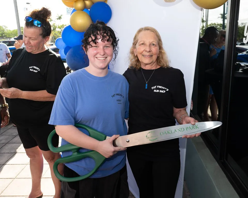 Abby June, 22, the new owner of Fat Freddy's Sub Shop, and Catherine Cerniglia stand side-by-side at the Oakland Park ribbon-cutting holding an oversized pair of ceremonial scissors, with blue and gold balloons behind them.