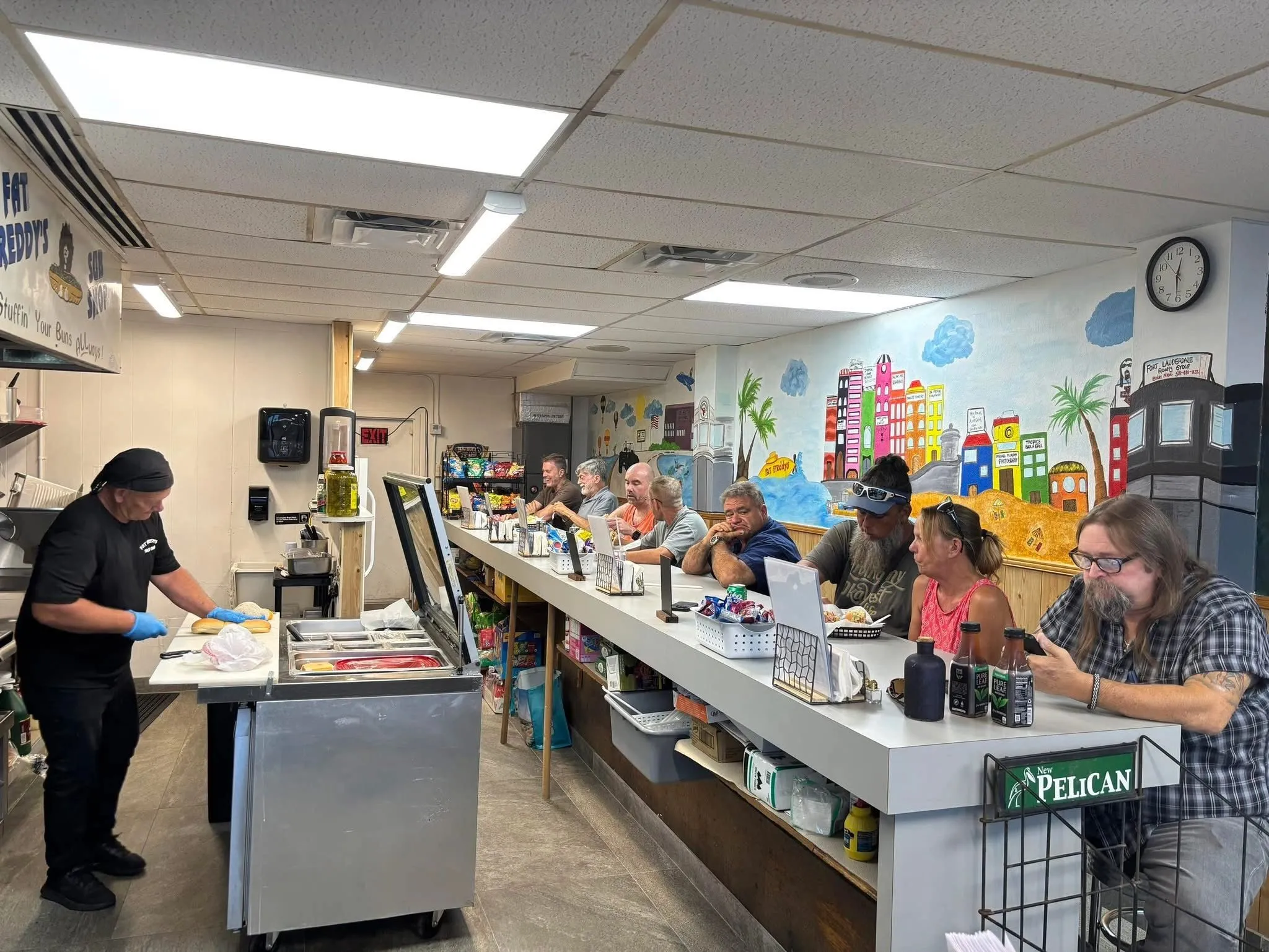Inside Fat Freddy's Sub Shop — counter, regulars, hand-painted Florida cityscape mural